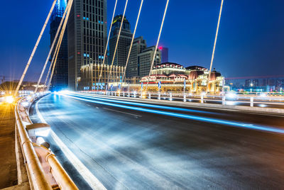 Light trails on city street by buildings against sky at dusk