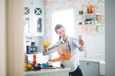 Multi-tasking mother food blogging while carrying daughter in kitchen at home