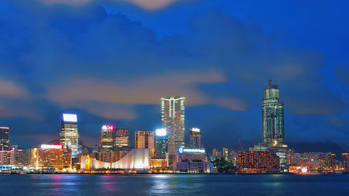 Illuminated cityscape in front of river against cloudy sky at dusk