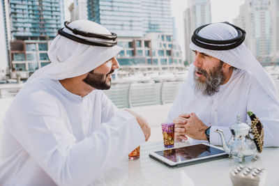 Men wearing dish dash discussing while sitting at outdoor restaurant in city