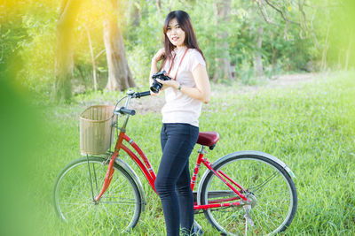 Portrait of woman with camera standing by bicycle on field against trees