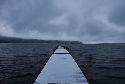 Snow covered pier amidst sea against cloudy sky