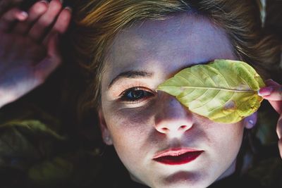 Close-up portrait of young woman holding leaf on eye while lying outdoors
