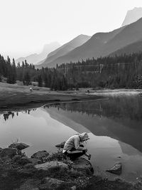 Man crouching at lakeshore against mountains