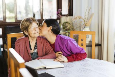 Caregiver kissing senior woman sitting at table in home