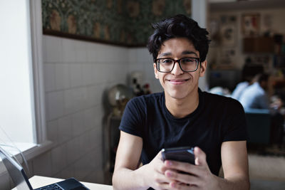 Portrait of smiling young man using social media on mobile phone while studying at home