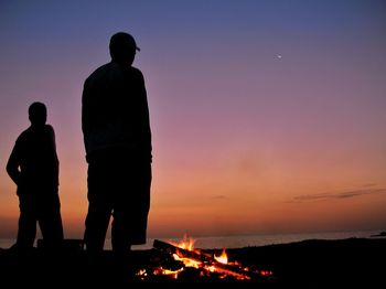Silhouette men standing by bonfire at beach during sunset