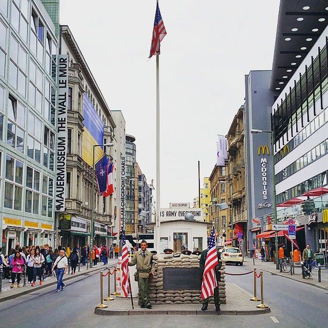 building exterior, architecture, built structure, city, street, city life, large group of people, person, flag, men, walking, road, city street, patriotism, clear sky, lifestyles, building, american flag, national flag