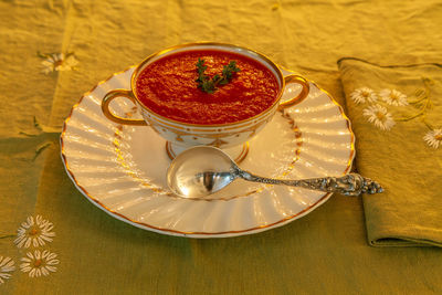 Tomato soup in a white and gold bowl on an embroidered green placemat.