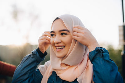 Smiling young muslim woman adjusting hijab against clear sky