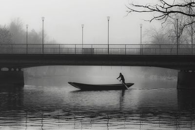 Silhouette man sailing boat on lake against bridge