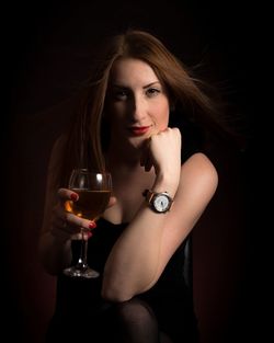 Portrait of young woman holding wineglass while sitting against black background