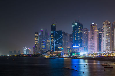 Illuminated buildings by sea against sky at night