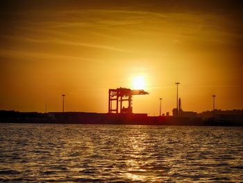 Silhouette factory by sea against sky during sunset
