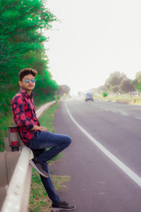 Portrait of young man sitting on road against sky and cars 