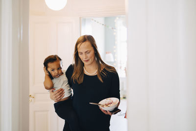 Mother with bowl carrying daughter talking on smart phone at home