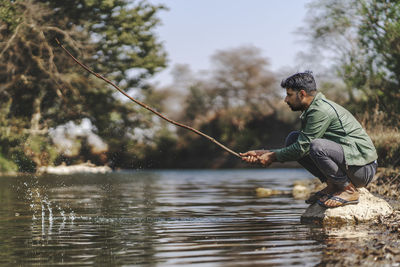 Man holding stick while crouching on rock in lake