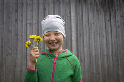 Portrait of a smiling girl holding flower while standing against wooden wall