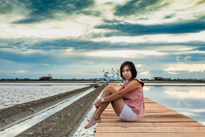 Portrait of mid adult woman sitting on pier over lake against cloudy sky during sunset