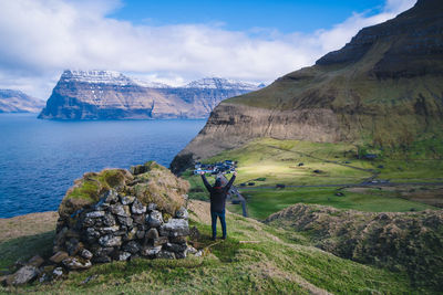 Man with arms raised standing on field by mountains against sky