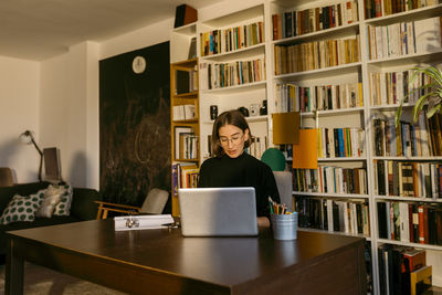 Female freelancer working on laptop sitting against bookshelf at home