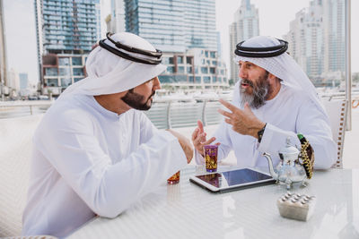 Men wearing dish dash discussing while sitting at outdoor restaurant in city