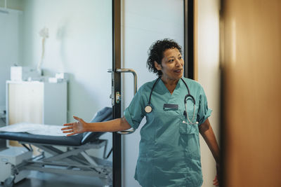 Female doctor wearing scrubs gesturing at doorway in hospital