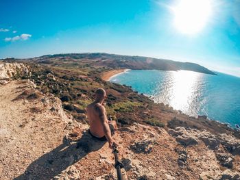 Rear view of man taking selfie sitting on rock by sea against sky