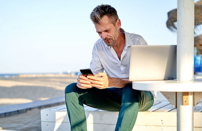 Middle aged male manager in smart casual clothes using laptop and browsing on smartphone while sitting on bench and working remotely from beach on summer day