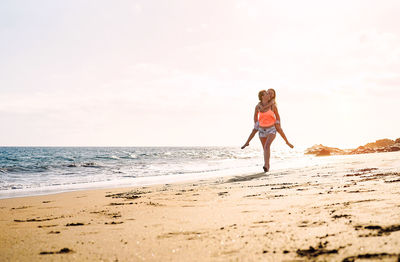 Woman carrying girl on back at beach against sky during sunset