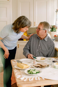 Mature female caregiver talking with retired senior man sitting at table for breakfast in nursing home