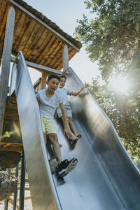 Brothers sliding on slide in public park on sunny day