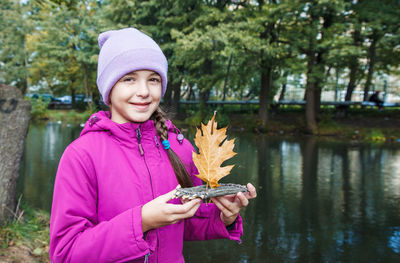 Portrait of smiling girl holding autumn leaf at lake