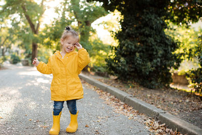 Pretty funny kid girl wear yellow raincoat, rubber boots walk in park over fallen leaves 