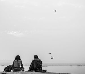 Rear view of men sitting by river against sky