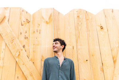From below young smiling hispanic man in black shirt with curly hair looking away while standing near lumber wall