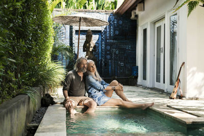 Senior man taking selfie at poolside while his wife is playing the guitar