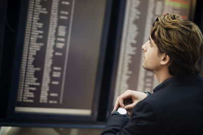 Businessman adjusting wrist watch while reading arrival departure board at airport