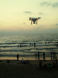 Drone flying over people at beach against sky during sunset