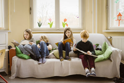 Full length of girls and boy reading books while sitting on sofa in classroom