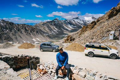 High angle view of man sitting on stone wall against cars and mountains