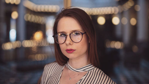 Portrait of young woman wearing eyeglasses against illuminated lights in city