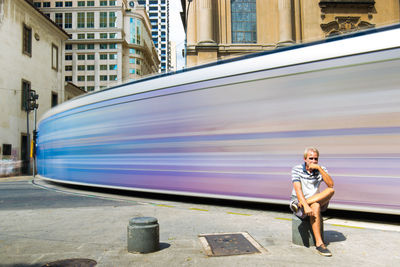 Full length of woman sitting by blurred motion of train in city