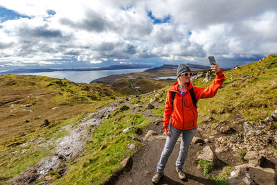 Woman taking selfie while standing on rock against cloudy sky