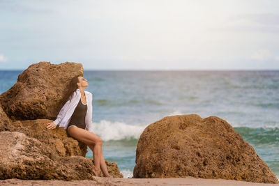 Rear view of woman sitting on rock at beach against sky