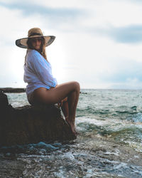 Young woman sitting on rock by sea against sky