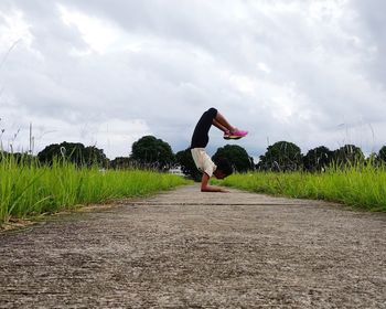 Side view of woman practicing handstand on footpath against cloudy sky