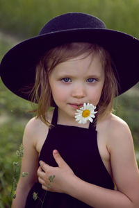 Child girl in a black hat and dress stands in a field with a camomile in her mouth in summer