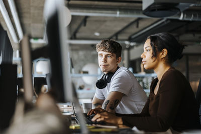 Young male computer programmer sitting next to female colleague working on computer at desk in office