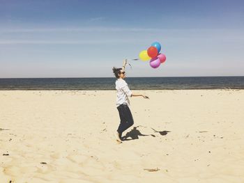 Side view of happy woman holding colorful balloons while walking on sand at beach during sunny day